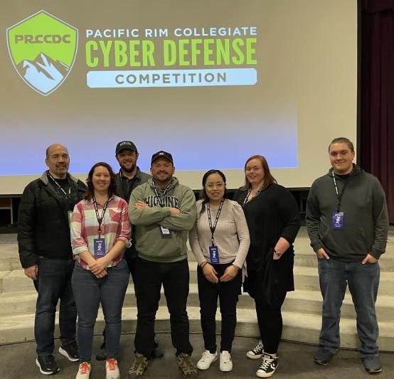 Picture of Cyber Competition Club members standing for a group picture in front of a backdrop that read "PRCCDC Pacific Rim Collegiate Cyber Defense Competition"